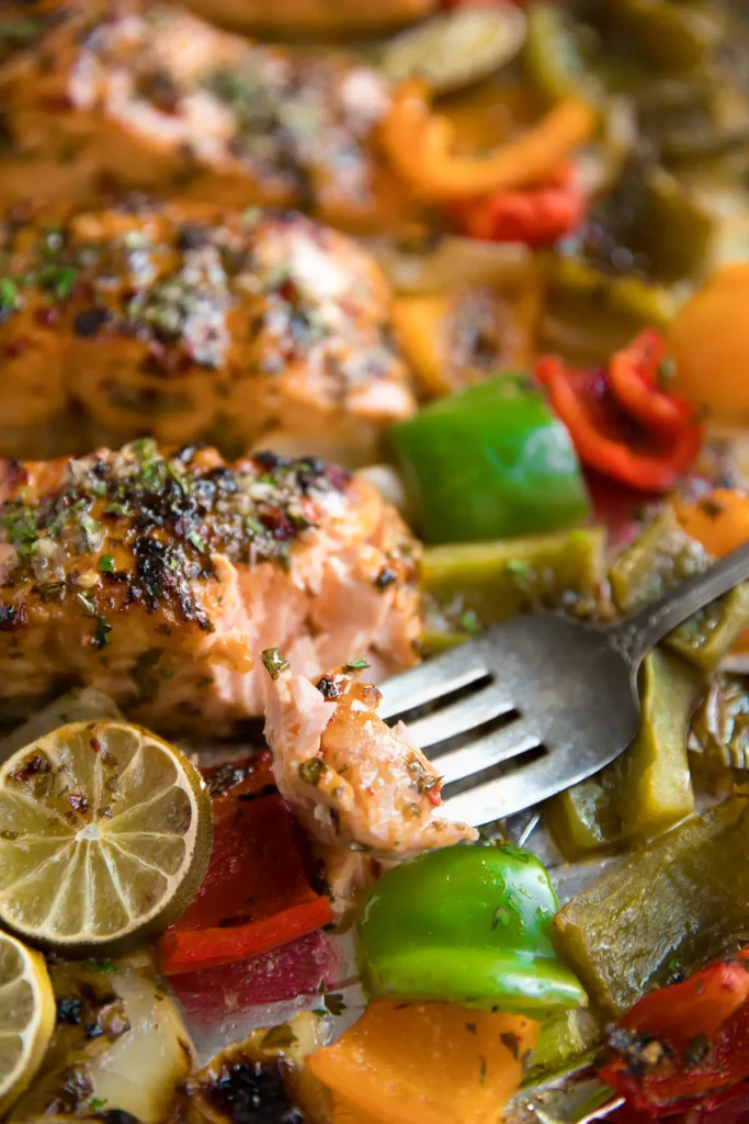 salmon baked on a sheet pan surrounded by colorful peppers with a fork taking a bite full out of the salmon