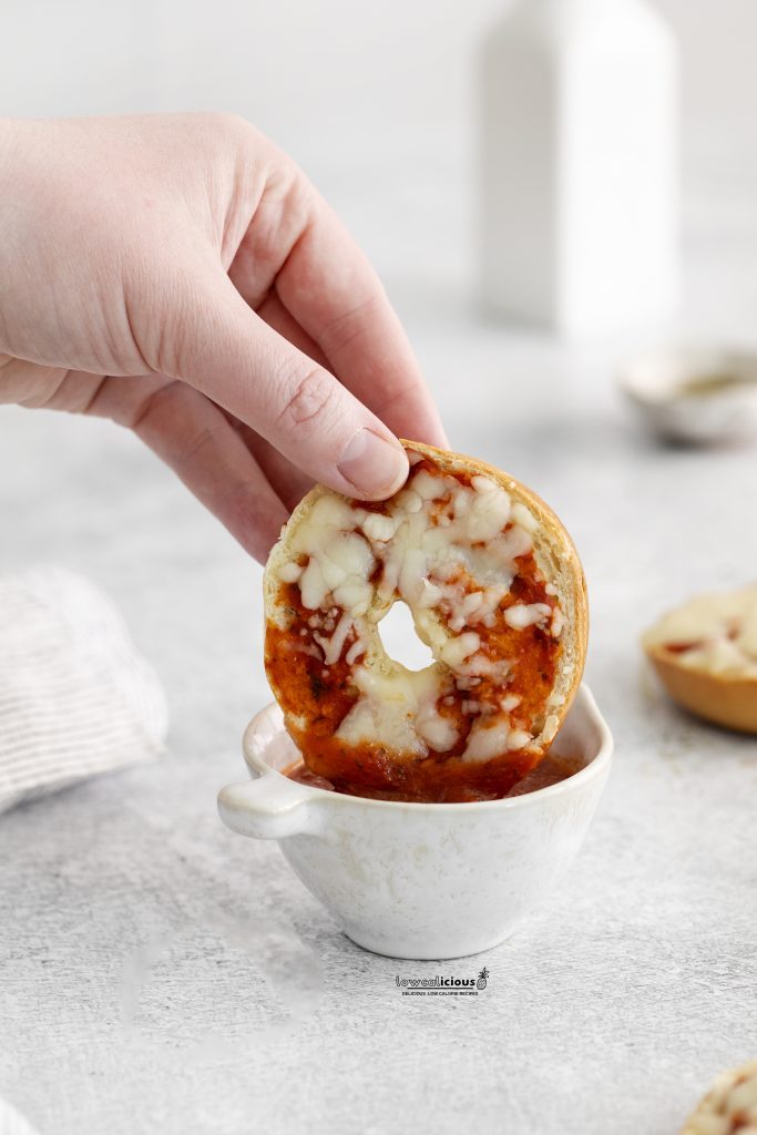 straight on shot of a hand dipping a low calorie pizza bagel in a small bowl of marinara sauce