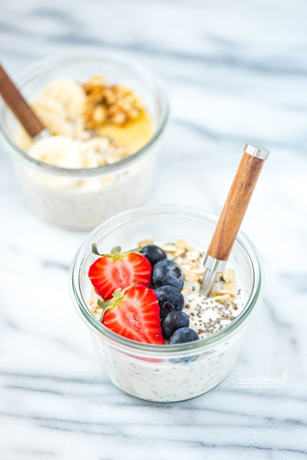 a bowl of overnight oats in a glass Weck jar with a wood handle spoon sticking out. the overnight oats are topped with a fresh strawberry that's been sliced in half, fresh blueberries, chia seeds, and some raw oats. In the background is a bowl of oats topped with banana slices, chopped walnuts, and honey.