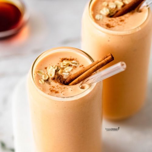two tall clear glasses filled with a Healthy Pumpkin Smoothie Recipe garnished with ground cinnamon, a few oats, a cinnamon stick, and a clear glass straw
