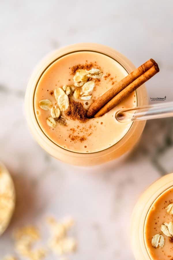 overhead shot of a healthy pumpkin smoothie in a clear glass garnished with raw oats, ground cinnamon, a cinnamon stick, and a clear glass straw