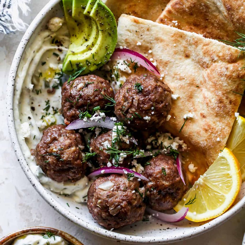 cooked Greek Meatballs in a white bowl garnished with lemon slices, pita bread, sliced red onion, avocado, and fresh herbs