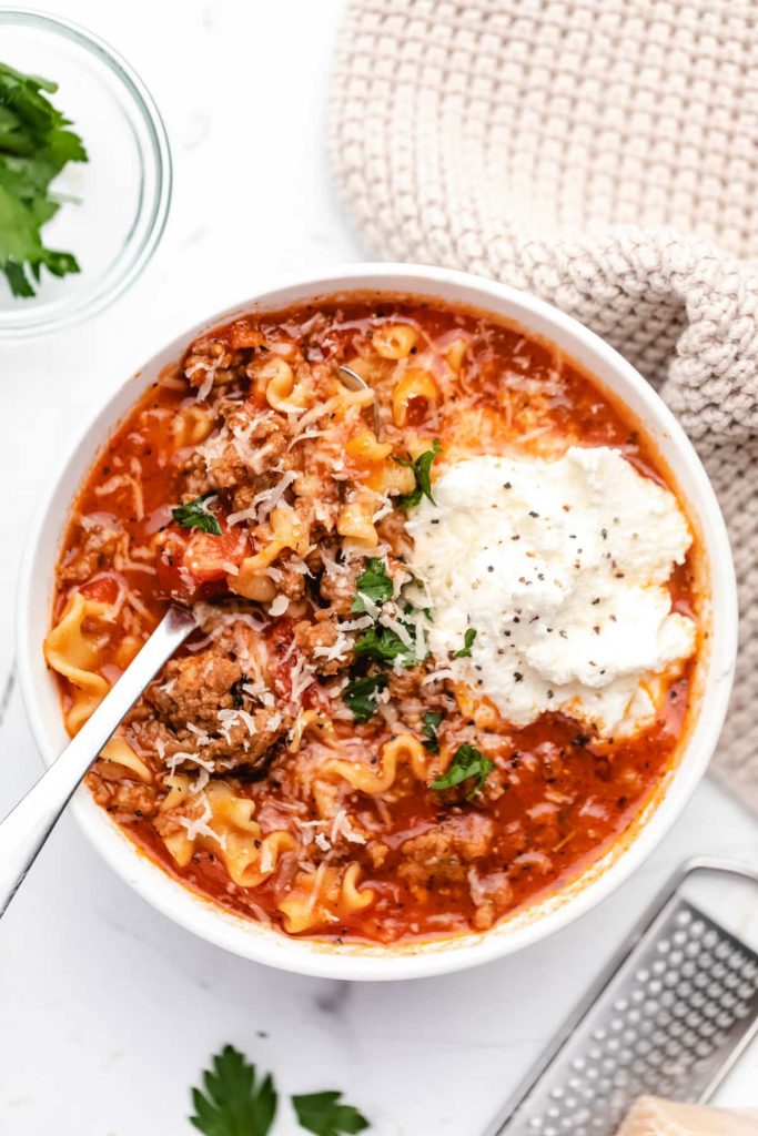 overhead shot of a bowl of Lasagna Soup in a white bowl with a spoon garnished with ricotta cheese and fresh parsley