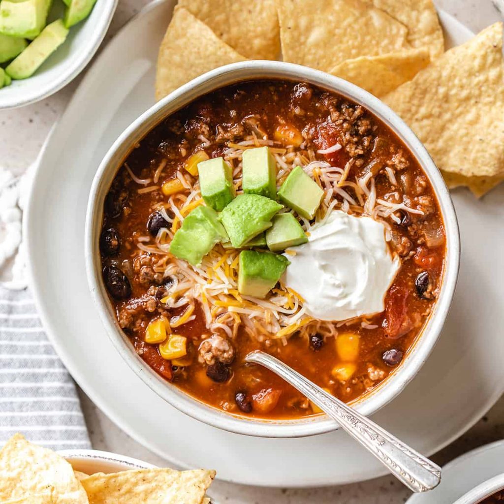 overhead shot of Slow Cooker Taco Soup in a white bowl with a silver spoon topped with diced avocado, shredded cheese, and a dollop of sour cream