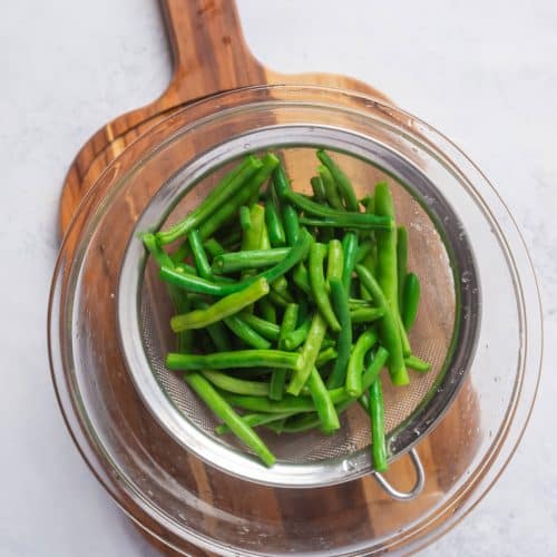 overhead shot of blanched green beans in a mesh strainer over a glass bowl on top of a brown cutting board to show the last step in how to blanch green beans