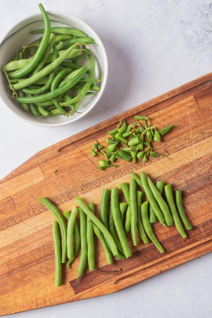 overhead shot of timed green beans on a brown wood cutting board with a white bowl of untrimmed green beans next to it to show the ingredients for how to blanch green beans