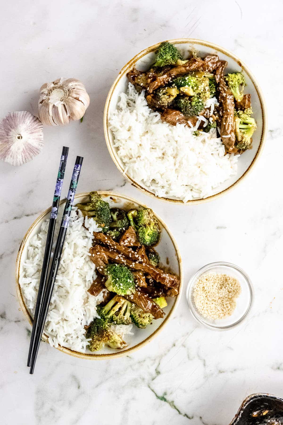 overhead shot of a prepared healthy beef and broccoli stir fry recipe plated in two bowls with white rice with chop sticks resting on one of the bowls