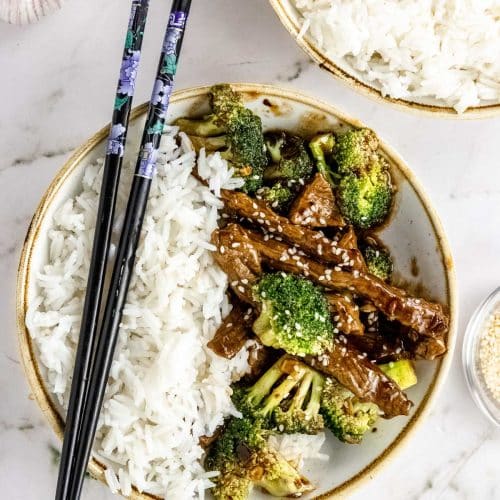overhead shot of the prepared healthy beef and broccoli stir fry recipe plated in a small bowl with white rice. Black chop sticks with purple flowers are resting on the side of the bowl.