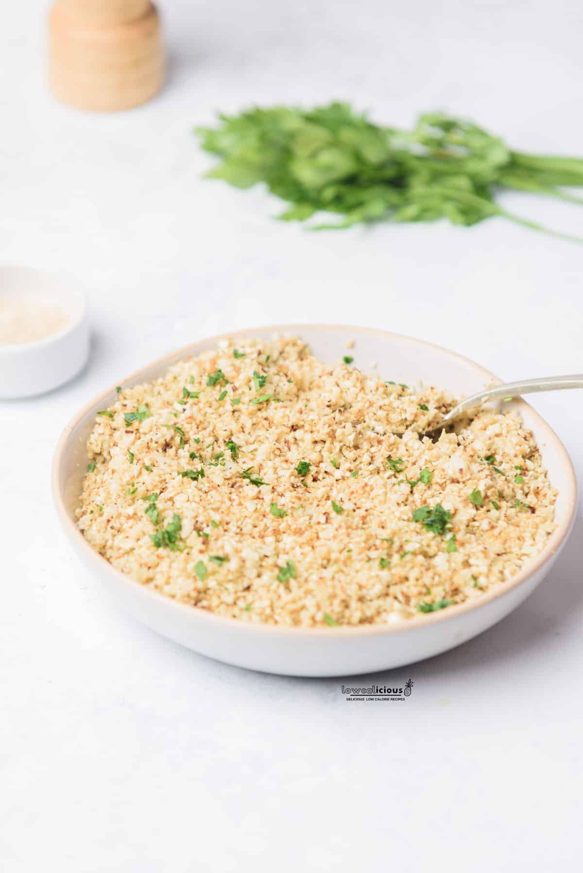 oven roasted cauliflower rice in a white serving bowl with a serving spoon in it, garnished with fresh parsley