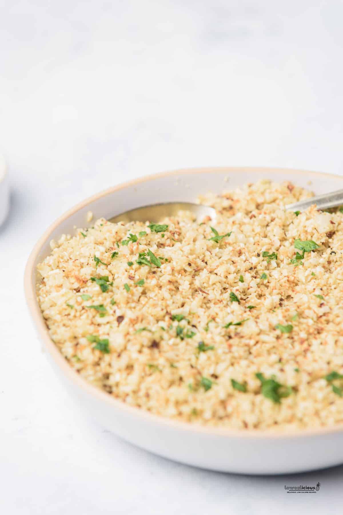roasted cauliflower rice in a white serving bowl garnished with chopped fresh parsley