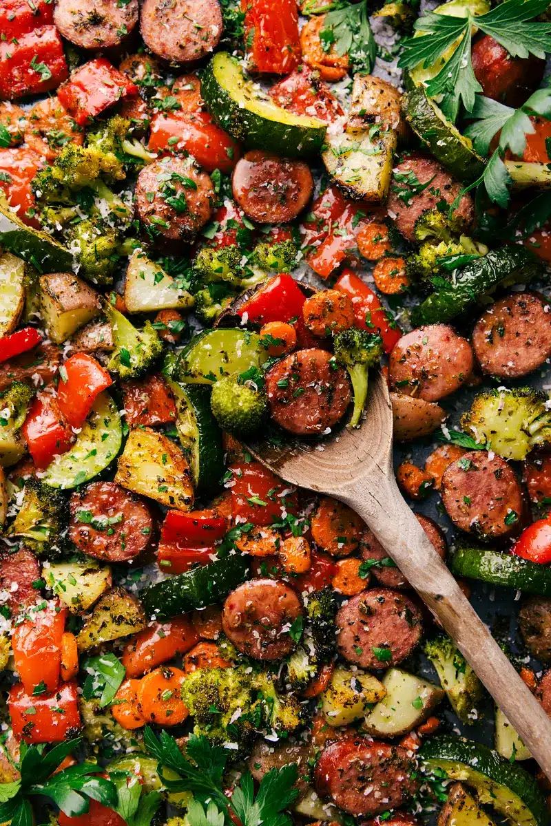 overhead shot of sheet pan sausage and veggies cooked with a wooden spoon stirring it - one of the recipes for this week's healthy weekly meal plan