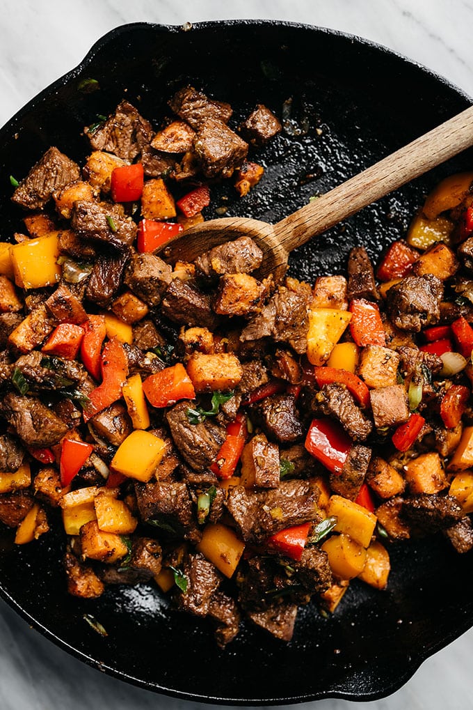 Whole30 Steak Bites with Sweet Potato and Peppers cooked in a cast iron skillet with a wooden spoon for this week's healthy weekly meal plan
