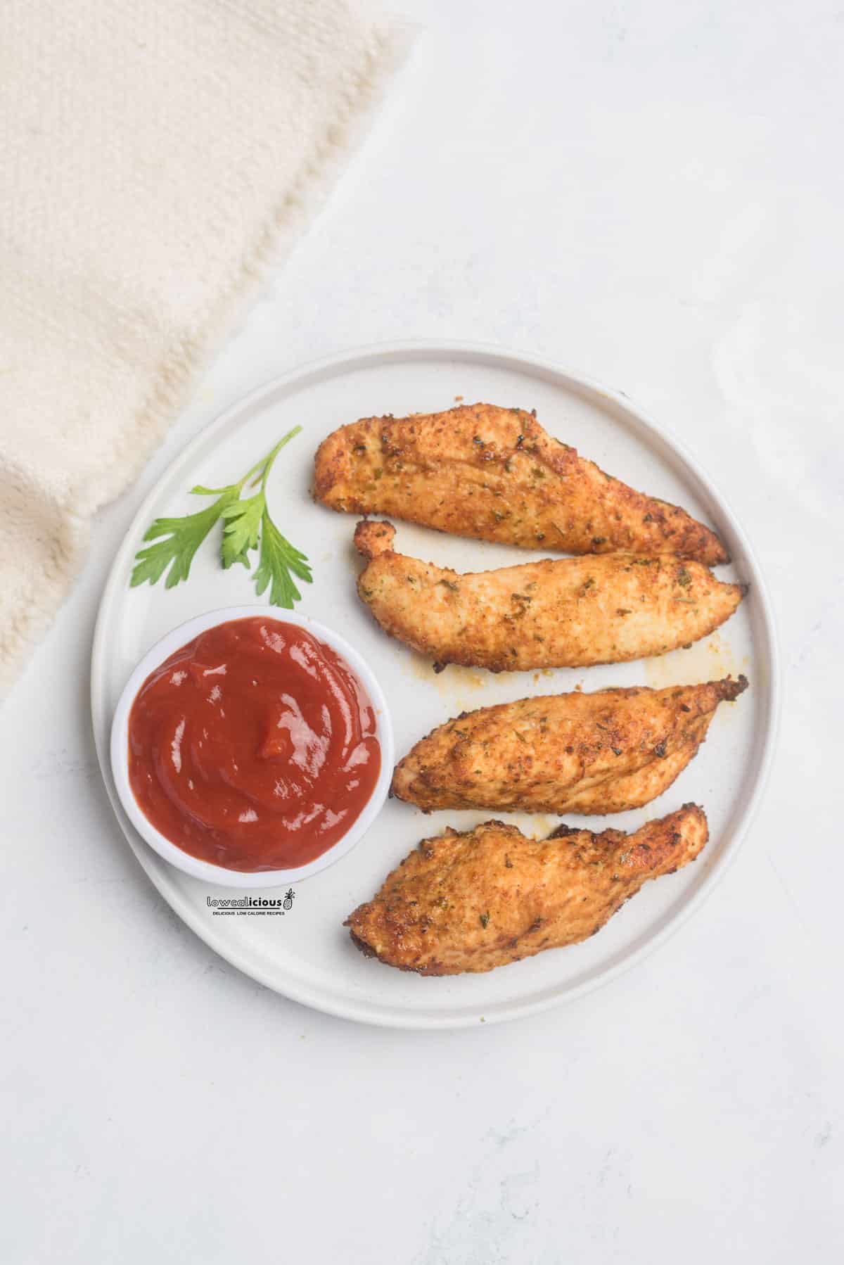 overhead shot of Healthy Air Fryer Chicken Tenders Recipe (No Breading) on a round white plate with a sprig of parsley and a small bowl of ketchup