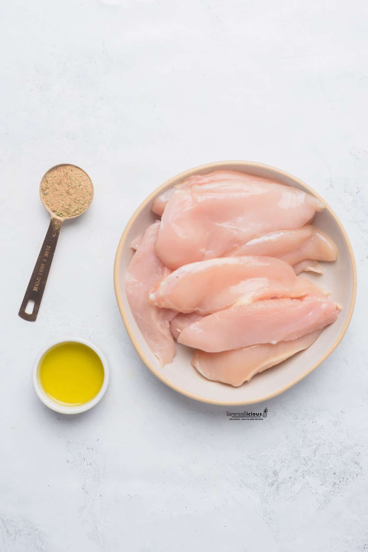 overhead shot of ingredients to make air fryer chicken tenders  - raw chicken tenders in a round white bowl, chicken seasoning in a measuring spoon, and a small bowl of oil