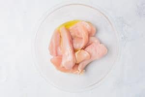 overhead shot of raw chicken tenders in a clear glass bowl with oil