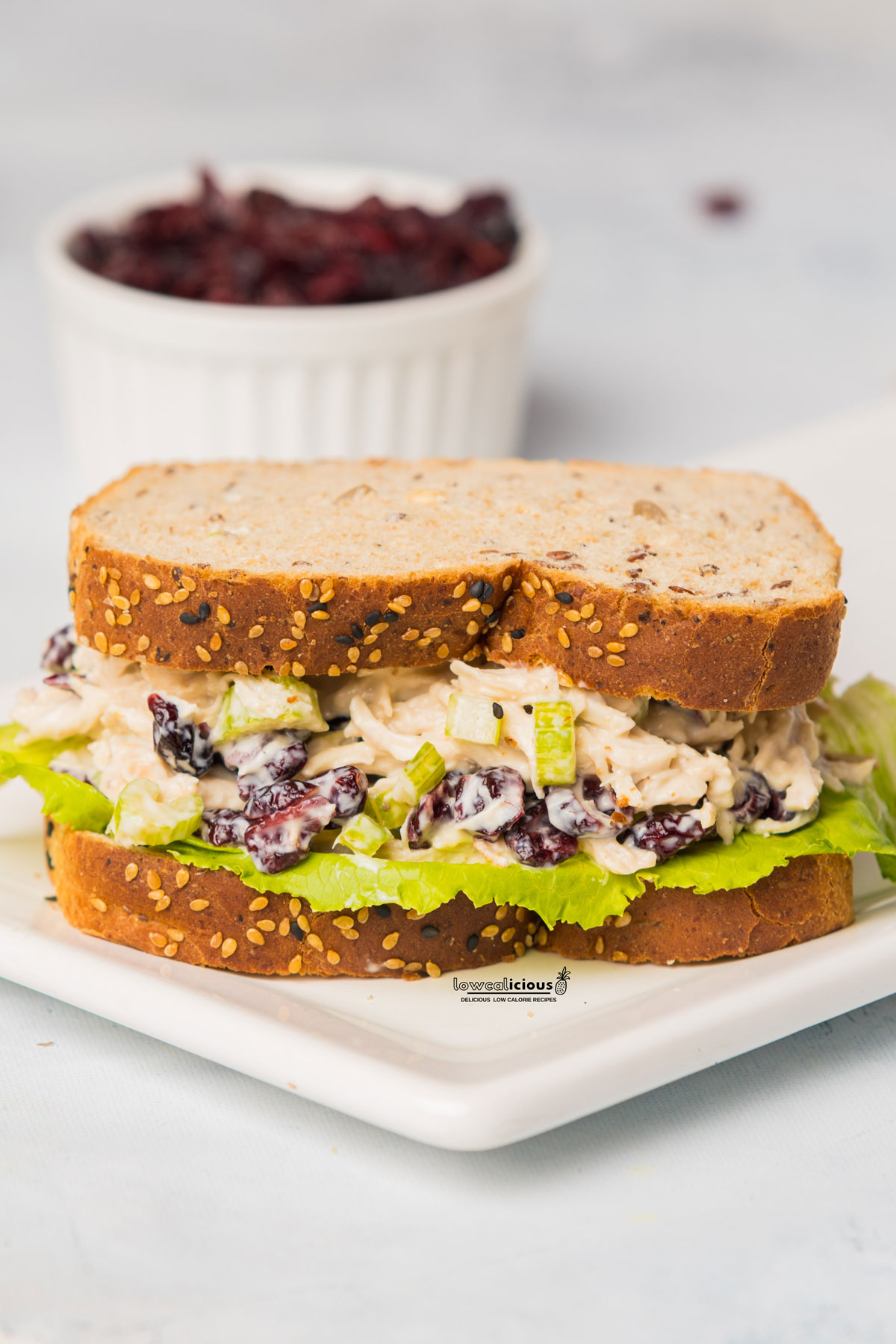 prepared Greek Yogurt Chicken Salad as a sandwich on two slices of bread with lettuce plated on a square white plate with a bowl of dried cranberries in the background