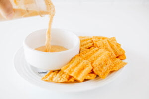 Viral TikTok Cottage Cheese Queso being poured into a small white bowl set on top of a round white plate with sun chips to serve