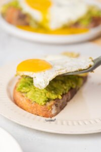 a spatula placing a cooked fried egg on top of a piece of avocado toast to make Pesto Eggs (Viral TikTok Recipe)