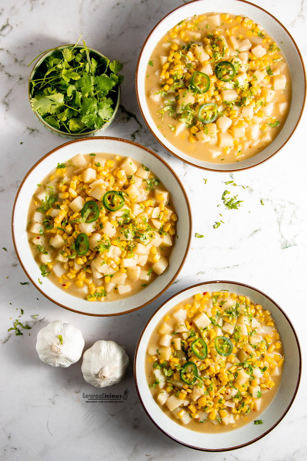 3 bowls of a cooked vegan corn chowder recipe with a small bowl of fresh cilantro and 2 heads of garlic nearby, ready to serve