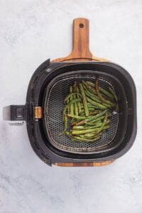 cooked green beans in a black air fryer basket on top of a wood cutting board