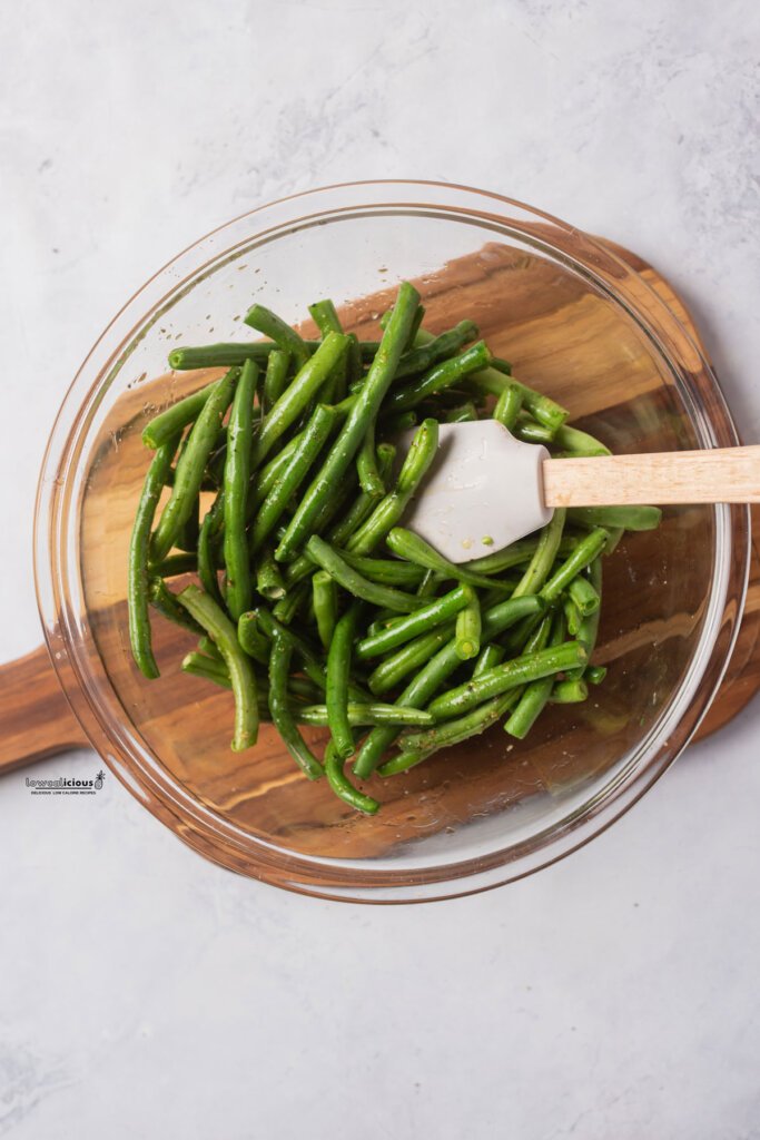 fresh green beans in a clear glass bowl with oil, garlic salt, and pepper that have been mixed with a white rubber spatula with a wood handle