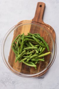 fresh green beans with black pepper and garlic salt in a clear glass bowl on top of a wood cutting board