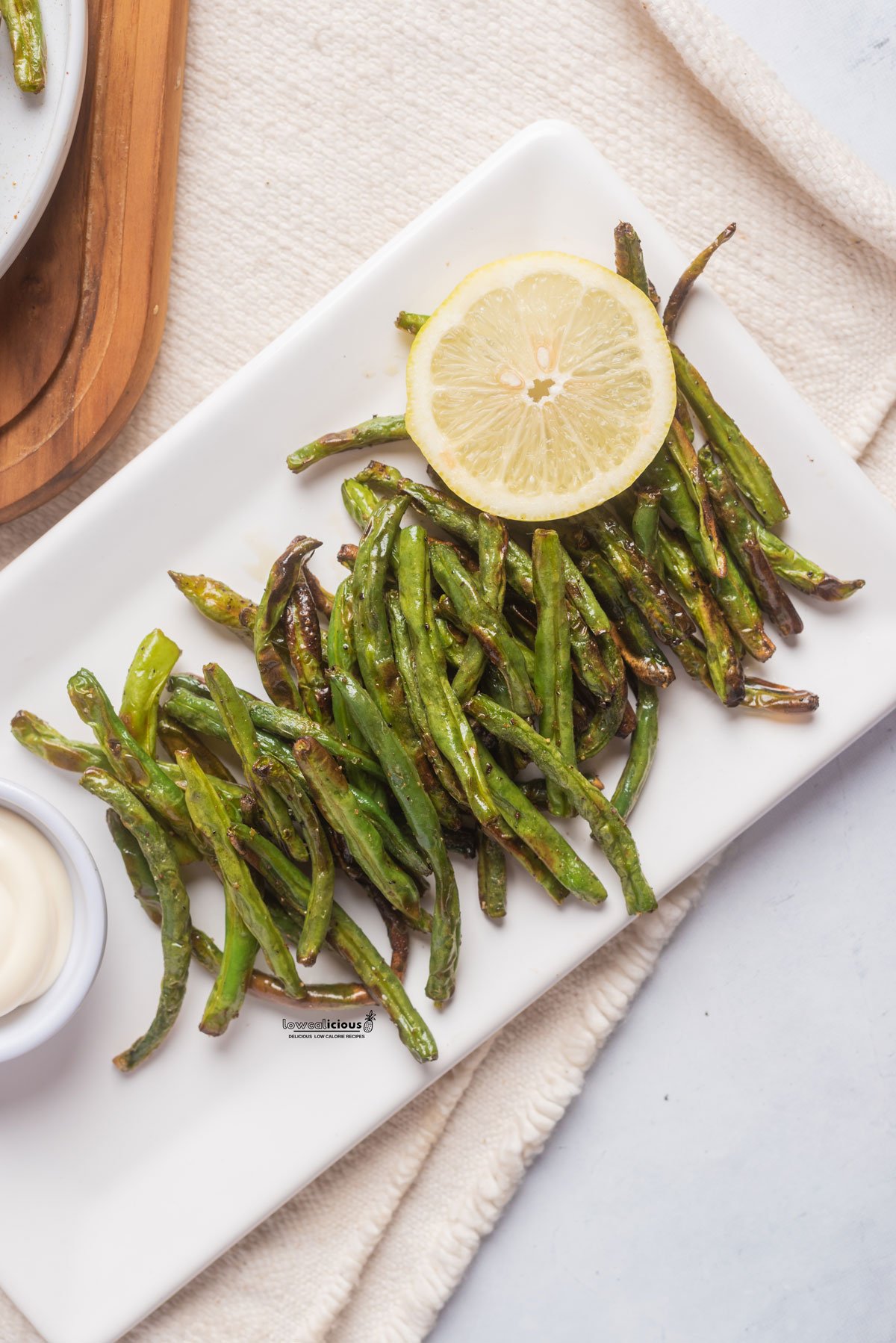cooked air fryer green beans on a white rectangle platter with a small bowl of aioli and a wheel of lemon