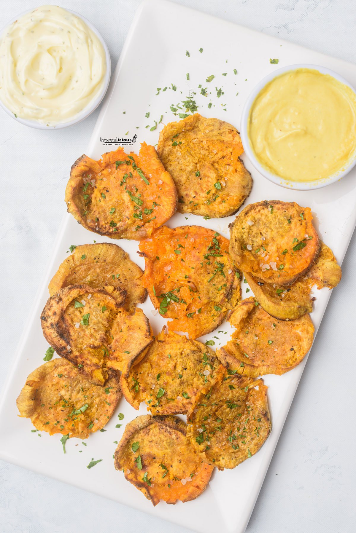 cooked Viral Smashed Sweet Potatoes made in an air fryer are plated on a white rectangular platter and topped with chopped fresh parsley alongside 2 bowls of dipping sauce.