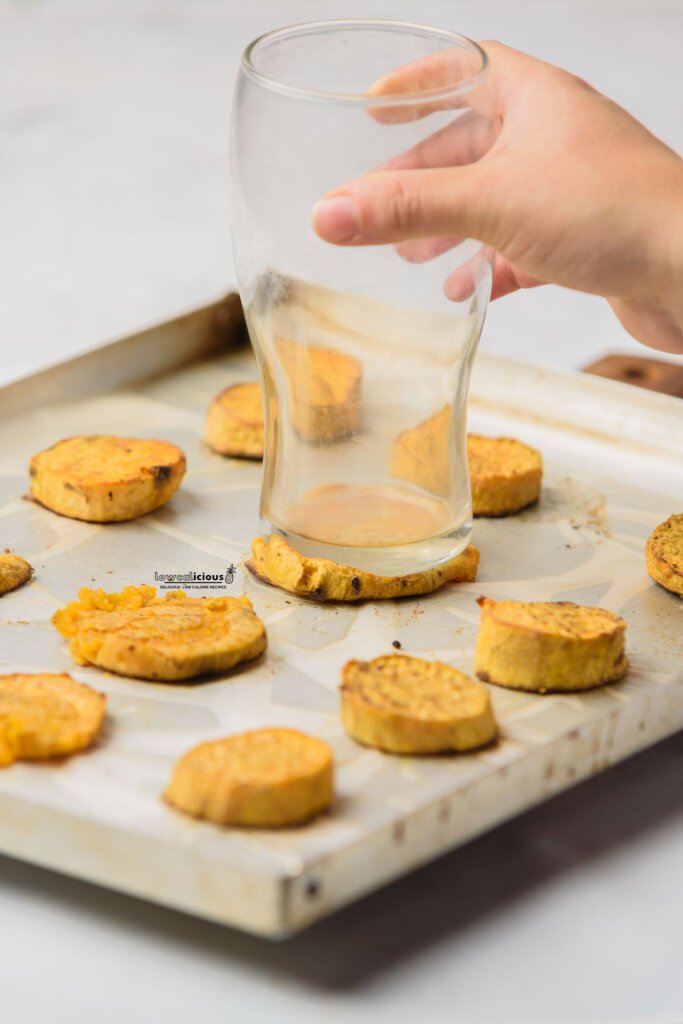 a hand holding a glass with a flat bottom to smash sweet potatoes on a parchment paper lined baking tray to make Viral Smashed Sweet Potatoes (Air Fryer Recipe)