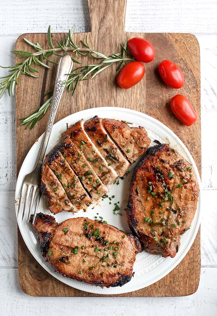 rosemary dijon pork chops on a round white plate on top of a brown wood cutting board with cherry tomatoes and a sprig of fresh rosemary