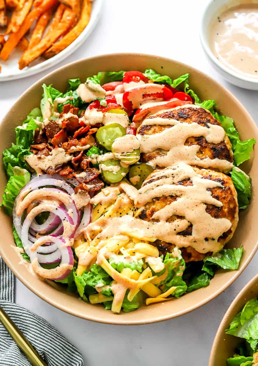 a green salad in a large tan bowl topped with lettuce, red onion, tomato, a burger, and special sauce to make a burger bowl