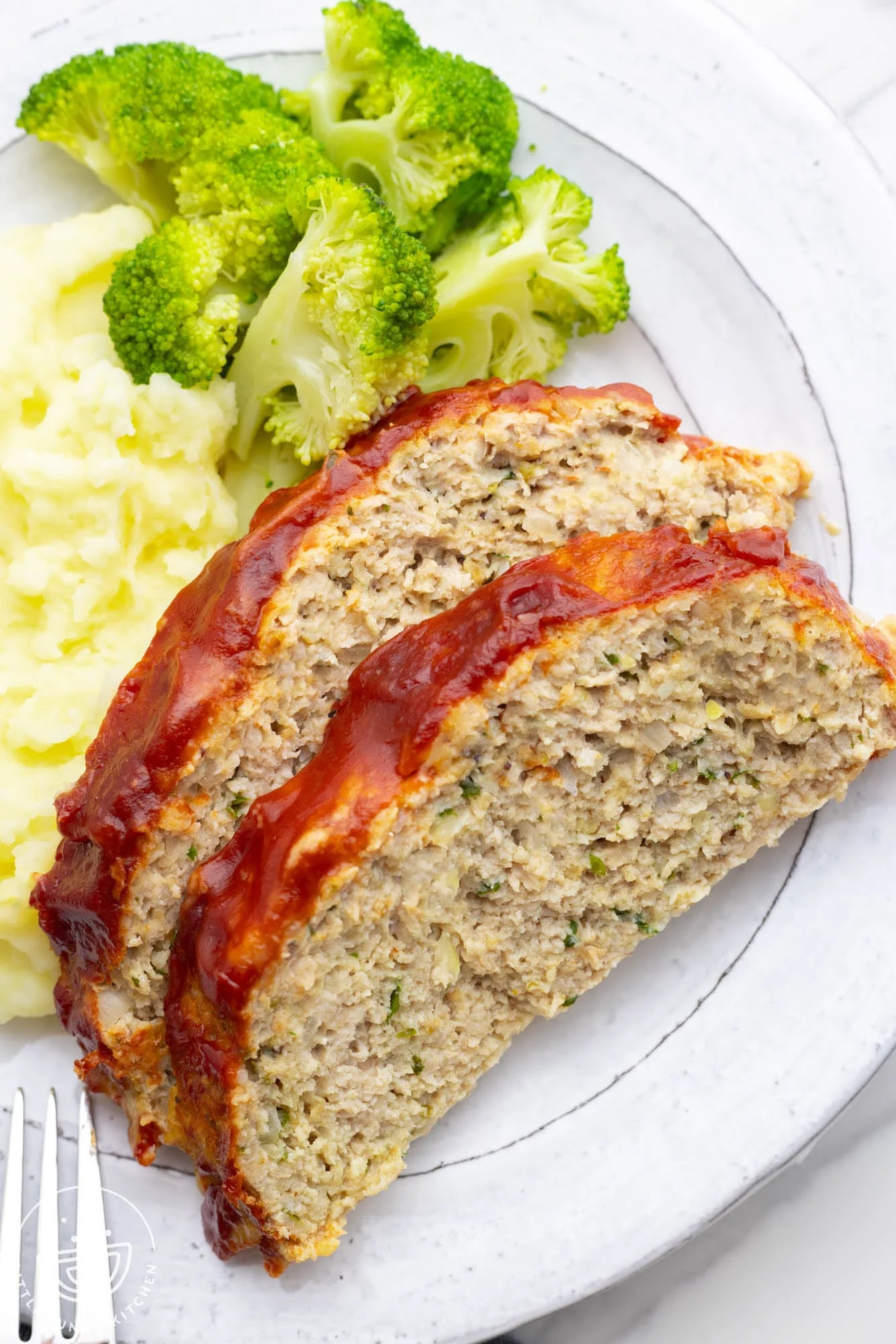 two slices of cooked chicken meatloaf with a red sauce topping on a round white plate with mashed potatoes and steamed broccoli