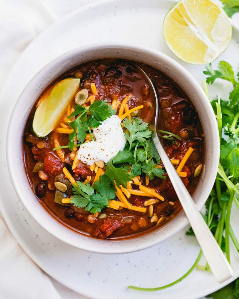 overhead shot of cooked instant pot vegetarian chili in a white bowl with a spoon on top of a white plate. The chili is garnished with a wedge of lime, fresh cilantro, and a dollop of sour cream for one of the recipes for this week's healthy weekly meal plan