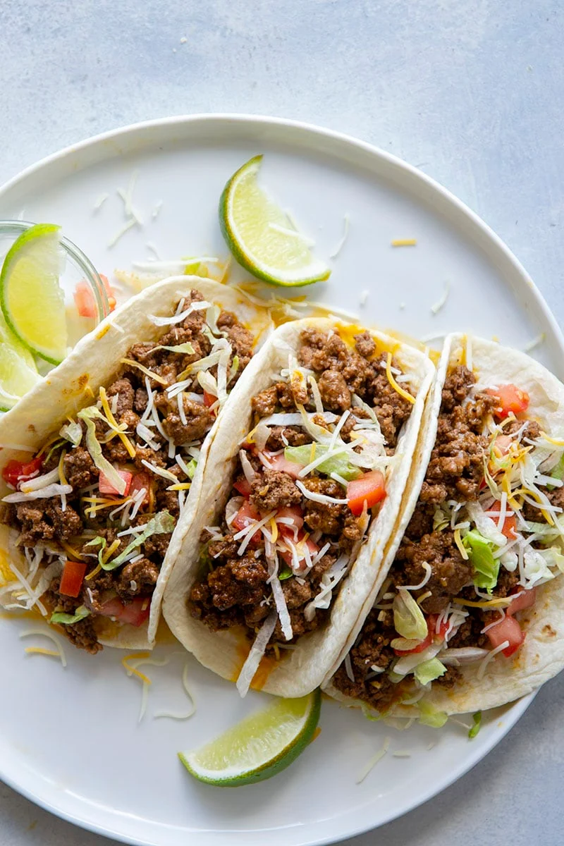 three keto tacos in low carb tortillas on a round white plate with lime wedges. The ground beef tacos are topped with shredded lettuce and diced tomato