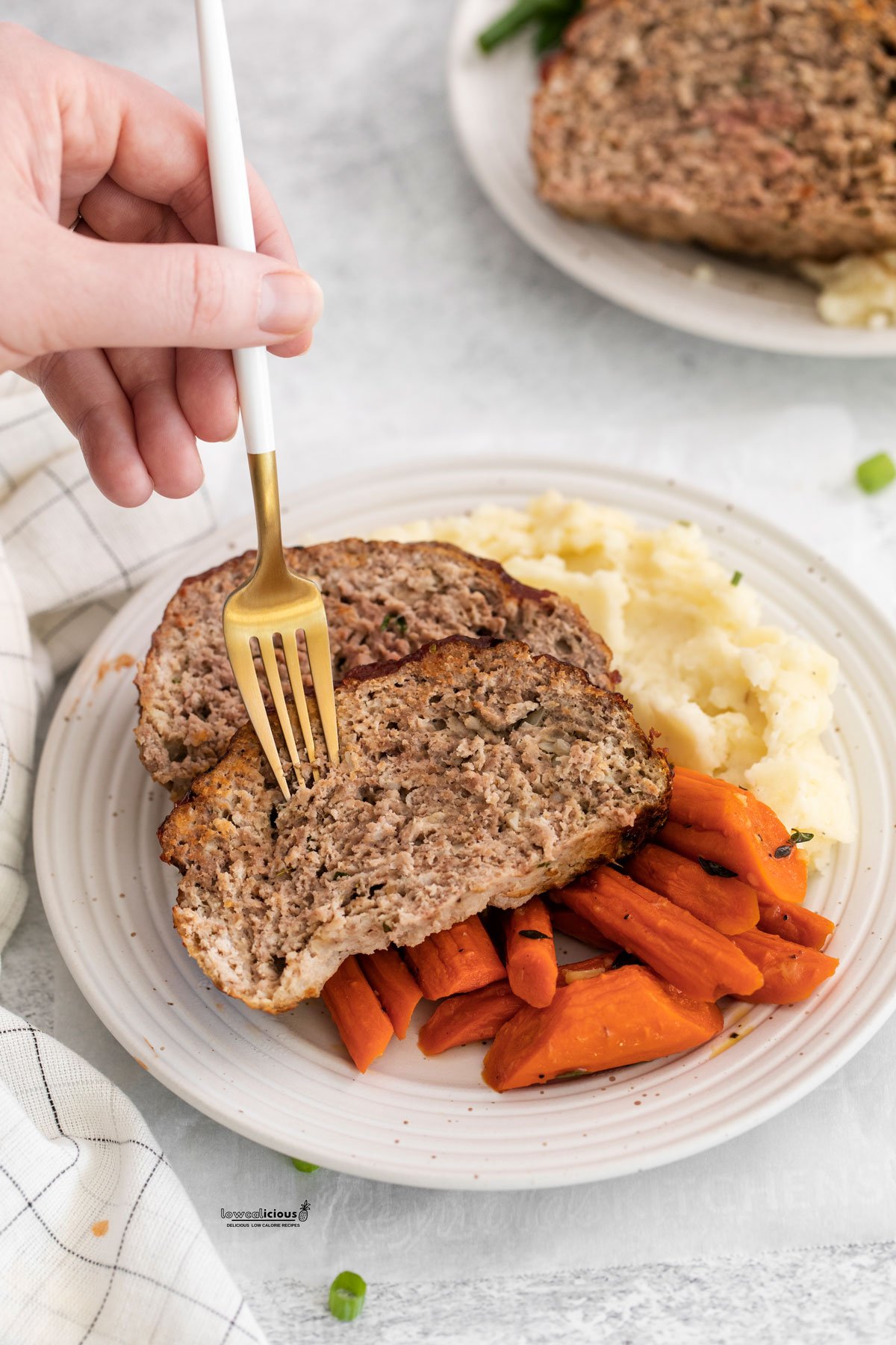 cooked Air Fryer Meatloaf on a white plate with mashed potatoes and carrots. A hand is holding a white and gold fork against a slice of meatloaf.